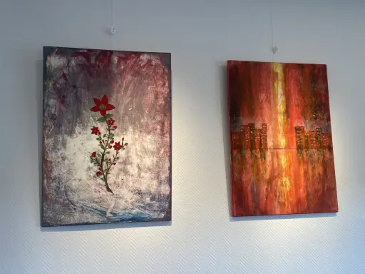 Två abstrakta målningar på väggen. Till vänster en röd blomma på en mörk bakgrund, en fin diy canvas. Till höger en stadssilhuett i varma färger.