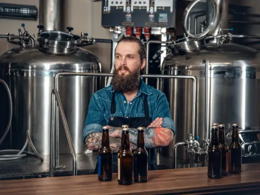 Un brasseur barbu pose devant une cuve chauffante, des bouteilles de bière sur le comptoir.