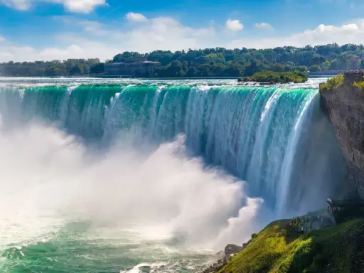 Potężny wodospad Niagara w całej okazałości. Woda spada kaskadami, tworząc mgłę. W tle drzewa i budynek.