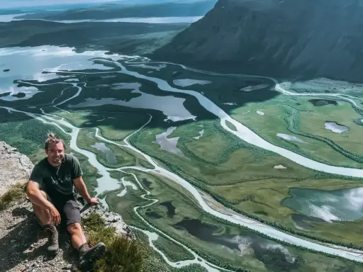 Man sitter på en klippa med en fantastisk utsikt över ett meandrande vattendrag och fjällandskap i Sarek. Aktse till skierfe!