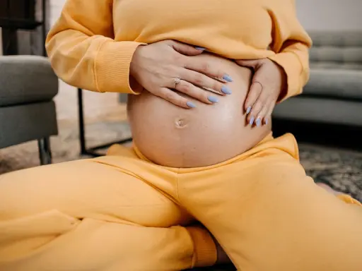 Schwangere Frau hält ihren Bauch, spürt den Druck im Unterleib. Gelber Jogginganzug, blaue Nägel.