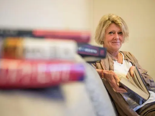 En kvinna med blont hår sitter med en bok i handen, omgiven av fler böcker. Hon ler mot kameran, som om hon delar en hemlighet från de fenomenala fruntimren på Grand Hotel.