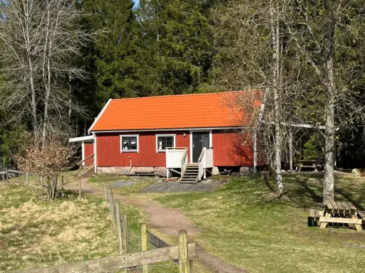 Rött torp med orange tak, omgivet av skog. En stig leder fram till torpet Freden, där picknickbord står redo för besökare.