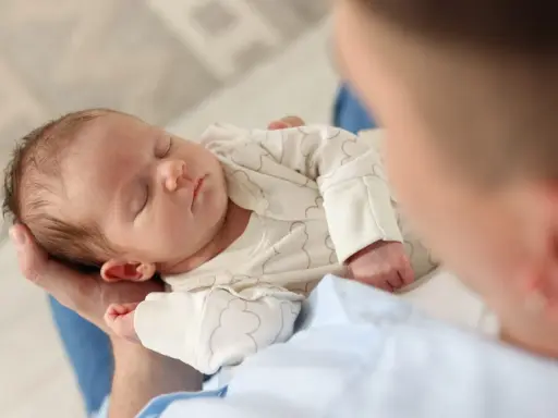 Un neonato dorme sereno tra le braccia di un genitore, cullato da suoni che ricordano i rumori bianchi, creando un'atmosfera di pace.