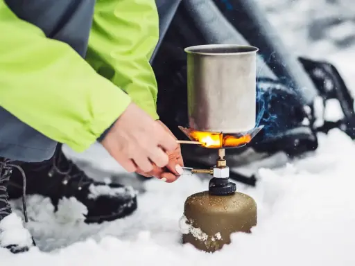 Tänd ett bensinkök i snön för att värma upp en kastrull. En person i en ljusgrön jacka och vandringskängor förbereder mat utomhus.