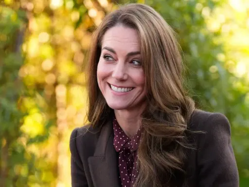 Kate Middleton sonríe con calidez, con su cabello castaño cayendo en cascada. El fondo bokeh de tonos otoñales complementa su elegante atuendo.