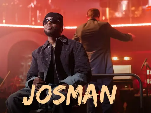 Josman, francuski rap artysta, siedzi na scenie z mikrofonem, w tle orkiestra i dyrygent.