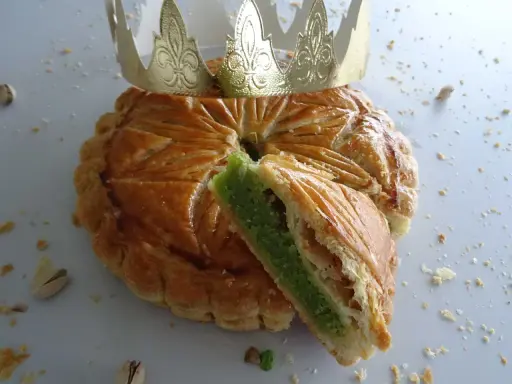 Galette des rois pistache dorée, une part coupée révèle sa garniture verte. Une couronne dorée trône sur ce délice.