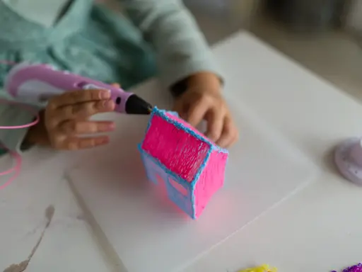 Dziecko tworzy różowy domek z niebieskimi detalami za pomocą różowego długopisu 3D.
