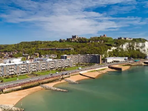 Vista aerea del porto di Dover con il castello in cima alla scogliera bianca. Edifici moderni e spiagge sabbiose si affacciano sul mare turchese.