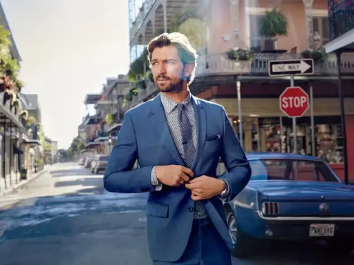 Man in a blue suit, perfect cocktail attire, stands on a street with a vintage car.