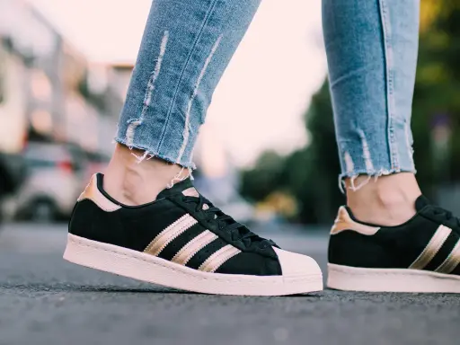 Nogawki jeansów i czarne **modele butów adidas z lat 2000.** ze złotymi paskami i różową podeszwą.