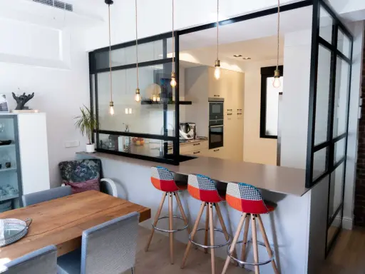 Tabique cocina semiabierta al salón con barra y taburetes coloridos. La cocina moderna se integra al salón a través de un panel de cristal y metal.
