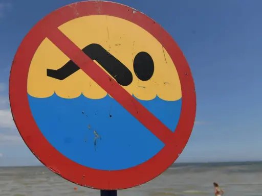 Znak zakazu kąpieli na plaży. Zamknięte kąpieliska nad morzem, mimo pięknej pogody i widocznej w oddali osoby w wodzie.