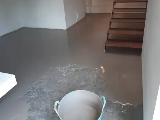 Pavimento continuo de hormigón gris recién aplicado, con una escalera de madera al fondo y un cubo con material.