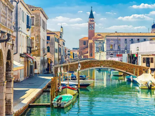 Un canal vénitien bordé de maisons colorées, avec des bateaux amarrés et un pont de briques. L'envie d'aller à Venise est forte.