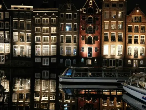 Edifici illuminati e riflessi sull'acqua, un battello turistico naviga tranquillo. Magica atmosfera di Amsterdam by night.