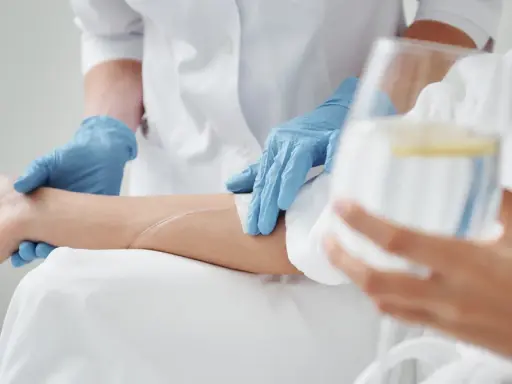 Pielęgniarka w niebieskich rękawiczkach zakłada wenflon do ręki pacjenta, przygotowując kroplówkę.