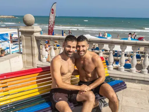 A happy couple enjoys a sunny day at the Sitges gay beach, sitting on a rainbow bench with the ocean in the background.