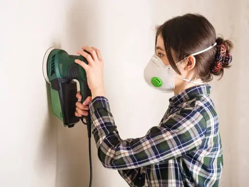 Mujer con mascarilla lijando paredes con una lijadora eléctrica.