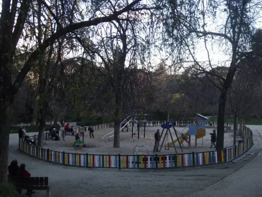 Parque de la Quinta de la Fuente del Berro con niños jugando en columpios y toboganes, rodeado de árboles y una valla colorida.