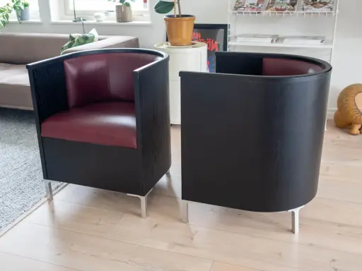 Två svarta fåtöljer med vinröda sitsar, designade av Mats Theselius, står på ett ljust trägolv.