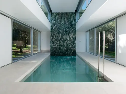 Piscina interior con pared de mármol verde veteado, rodeada de ventanales que dan a un jardín.