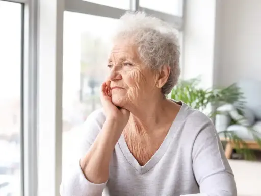 Anziana con mano sul mento, sguardo pensieroso verso la finestra. Potrebbe riflettere sulla pseudodemenza depressiva.