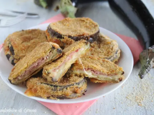 Cordon bleu di melanzane impanati e fritti, ripieni di prosciutto e formaggio filante, serviti su un piatto bianco con melanzane fresche sullo sfondo.