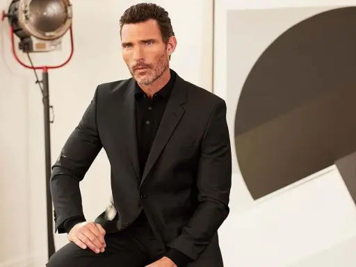 A man in a sharp black suit and polo shirt, a classic funeral outfit for men, poses with a thoughtful expression.