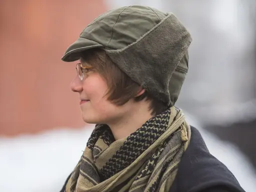 Osoba w okularach nosi zieloną czapkę polową z futrzanymi nausznikami i szalik w stylu keffiyeh.