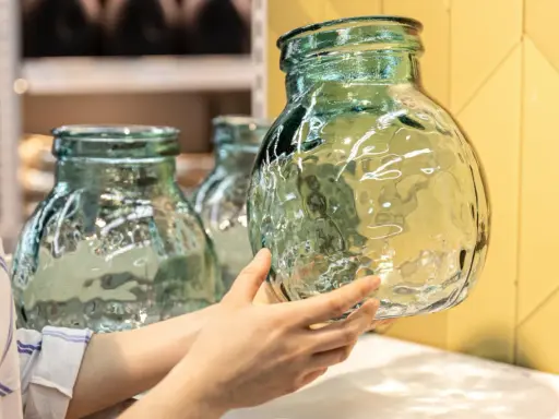 Une main examine un objet en verre texturé, vert pâle, aux reflets ondulés. D'autres objets similaires sont visibles en arrière-plan.