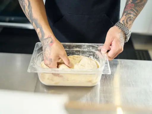 Ręce z tatuażami zagniatają ciasto w plastikowym pojemniku. To pierwszy krok, by dowiedzieć się, jak mrozić ciasto.