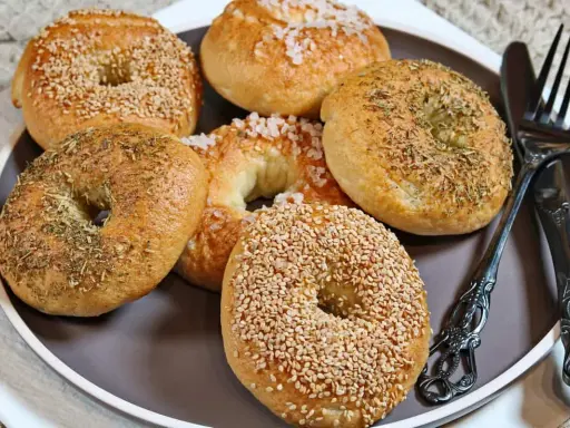 Sechs selbstgemachte Bagels auf einem Teller: zwei mit Sesam, zwei mit Kräutern und zwei mit grobem Salz bestreut. Ein Messer und eine Gabel liegen daneben.