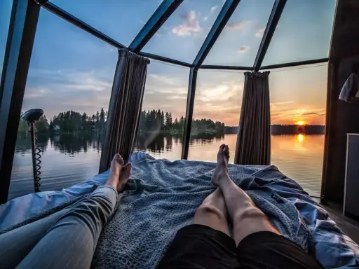 Två personer vilar i en glaskupa med utsikt över en sjö i Norrland vid solnedgången. En perfekt glampingupplevelse.
