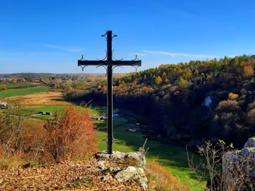 Czarny krzyż na skale góruje nad jesienną doliną Grzybowską. W oddali widać pola i lasy w barwach złota i czerwieni.