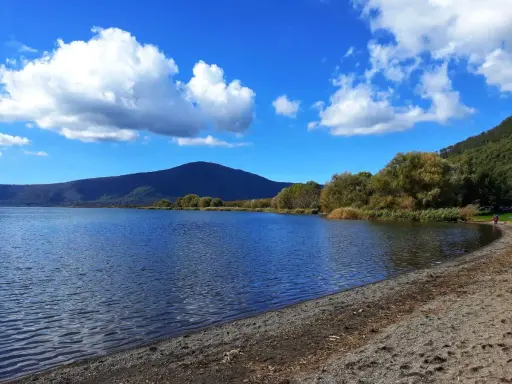 Spiaggia ghiaiosa e acque calme del lago di Vico, con montagne verdi e cielo azzurro punteggiato di nuvole bianche.