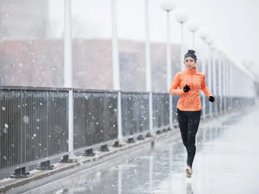 Donna corre sotto la pioggia su un ponte, con fiocchi di neve che cadono. Indossa abbigliamento sportivo arancione e nero.