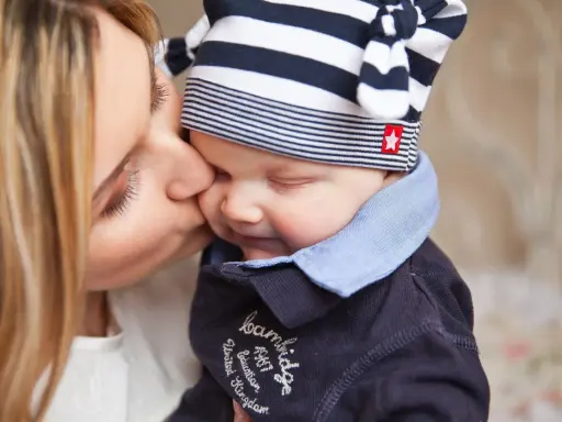 Delikatny pocałunek mamy, by obudzić dziecko. Uśmiechnięta twarz malucha w czapce w paski.