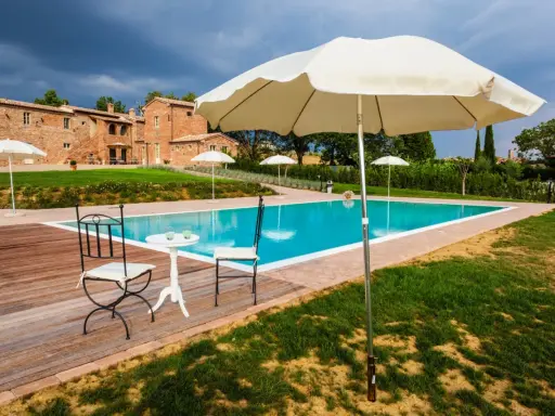 Piscina con vista su un casale in mattoni, perfetta per le tue vacanze in Toscana. Tavolino con sedie e ombrelloni pronti per rilassarsi.