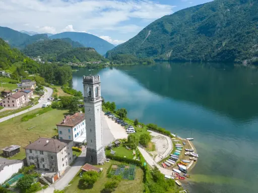 Torre campanaria affacciata sul lago di Corlo, con montagne verdi sullo sfondo e un villaggio pittoresco.