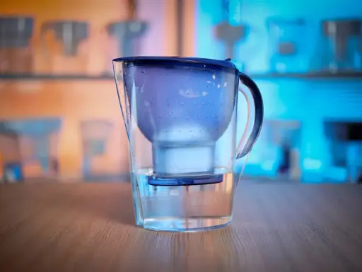 En vattenrenare för dricksvatten, en Brita-kanna, står på ett träbord. Bakgrunden visar fler liknande kannor i blått och orange ljus.