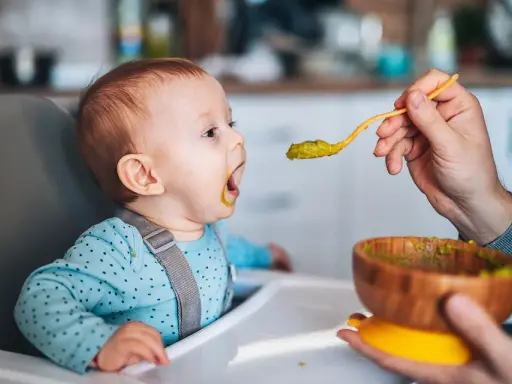 Ein Baby sitzt im Hochstuhl und bekommt von einer Hand mit einem Löffel Babybrei mit Fleisch gefüttert.