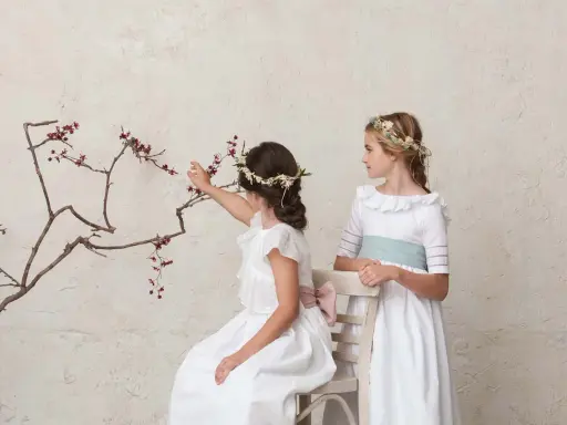 Dos niñas con vestidos blancos y coronas de flores, una sentada tocando una rama, la otra de pie. Peinados de trenza para niña de comunión.