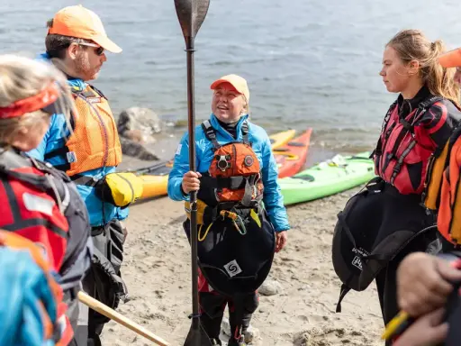 Grupp samlad på stranden, redo för paddling. En instruktör visar paddelteknik. Kajaker ligger i bakgrunden.