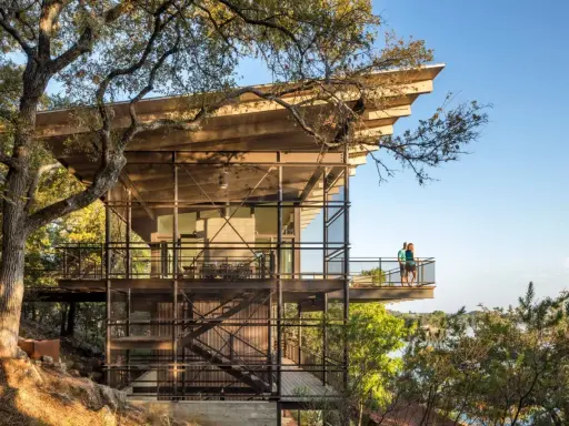 Maison moderne sur un terrain en pente raide, avec des balcons et des escaliers extérieurs en métal, nichée dans les arbres près d'un lac.
