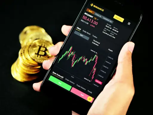 Ręka trzyma smartfon z wykresem kursu Bitcoina na platformie Binance. Obok stos złotych monet. Czy Binance czy Zonda? Decyzja należy do Ciebie.