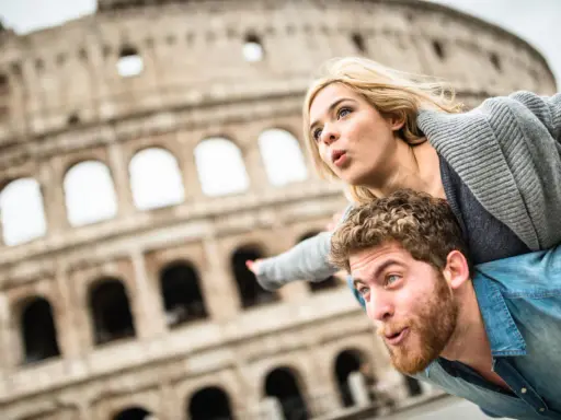 Coppia in una vacanza romantica a Roma, con il Colosseo sullo sfondo. Lui porta lei sulle spalle, entrambi con espressioni sorprese.