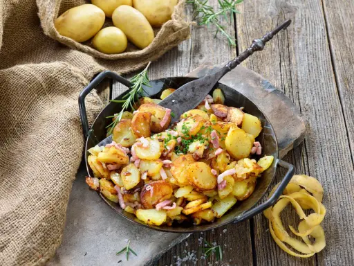 Bratkartoffeln, die vorher gekocht wurden, in einer Pfanne mit Speck und Kräutern garniert. Daneben liegen rohe Kartoffeln in einem Jutesack.