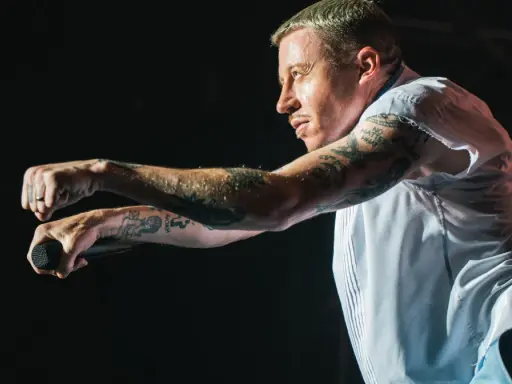 Macklemore na koncercie w Polsce, z mikrofonem w dłoni, w białej koszuli i z tatuażami na ramionach.
